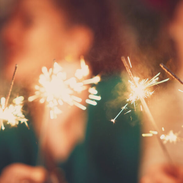 Des cierges magiques s’allument au premier plan d’une photo festive du Nouvel An avec plusieurs personnes qui rient en arrière-plan, paraissant floues.